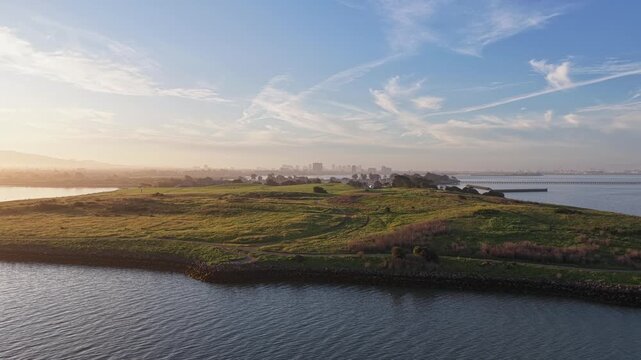 Aerial zoom out drone shot showing Cesar Chavez Park in Berkeley, California, with the calm waters of the bay and a clear blue sky during a beautiful hazy sunrise lighting the landscape. Filmed in 4k