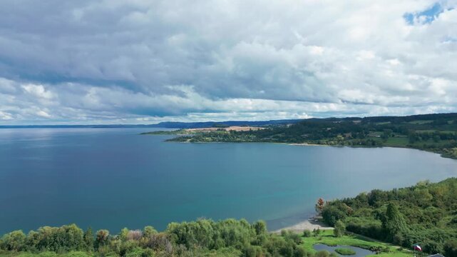 Aerial view of vast blue lake with forested shoreline and small town on distant peninsula under dramatic cloudy sky with moody light. Futrono, Chile