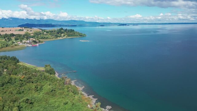Aerial view of lush forest peninsula and turquoise lake stretching to distant islands and mountain ranges under bright blue sky. Futrono, Chile
