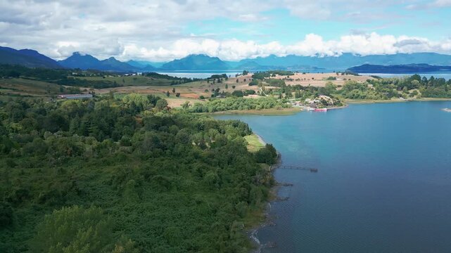 Panoramic pan over lush forest peninsula and golden farmland along vast lake shoreline with dramatic Andean mountain range in the background. Futrono, Chile