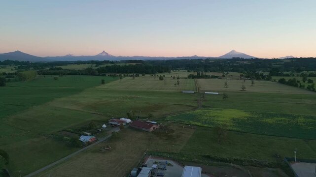 Backward flight over rural farmland and isolated farm buildings revealing vast Andean volcanic range glowing in soft pastel sunset light. Entre Lagos, Chile