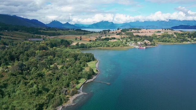 Tilting down shot over forested peninsula and calm blue lake revealing golden farmland and dramatic Andean mountain range in the distance. Futrono, Chile