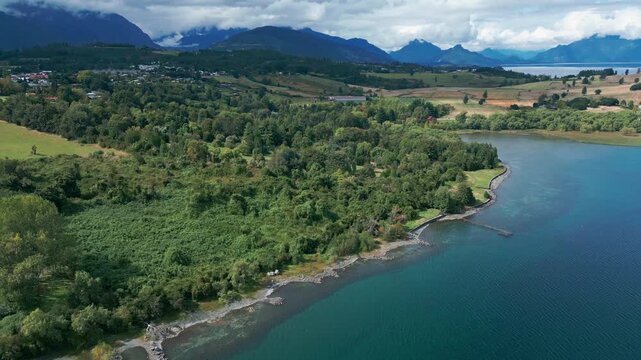 Reveal shot over lush forest shoreline unveiling turquoise lake, rolling farmland and dramatic cloud-covered Andean mountain range beyond. Futrono, Chile