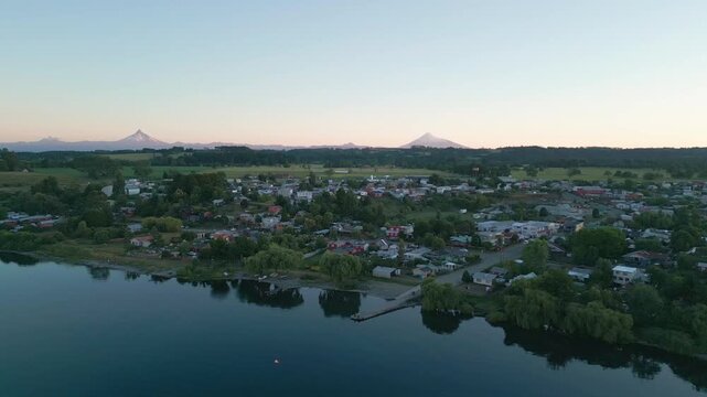 Backward flight over calm lake revealing small lakeside town with snow-capped Andean volcanoes and pastel sunset sky on the horizon. Entre Lagos, Chile