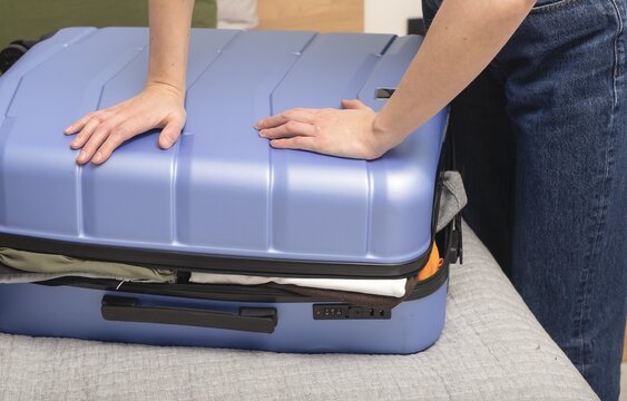 Person pressing suitcase lid to close overpacked luggage during travel preparation. Hands compress contents inside case. Concept of packing overload organization journey..