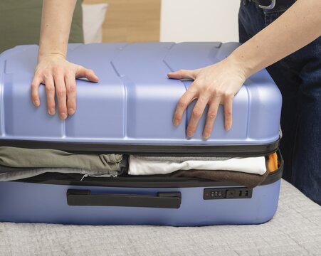 Woman pressing suitcase lid to close overfilled luggage during packing. Hands apply force to compress contents inside case. Concept of travel preparation overload..