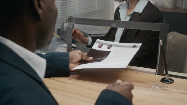 Medium close-up of middle-aged African American man in suit checking photos and application form before submitting passport for business visa to cheerful Caucasian female consulate official