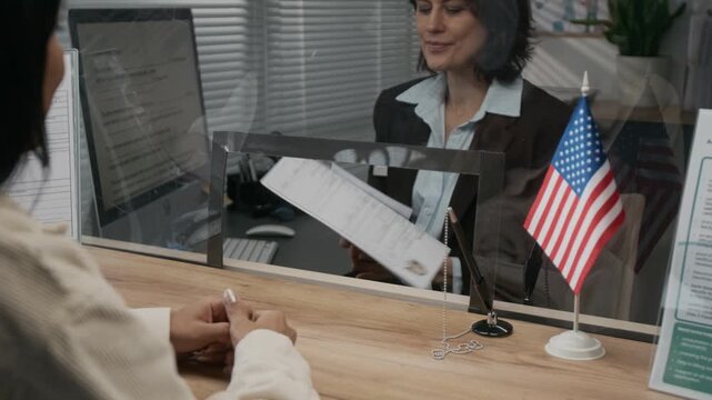 Over-shoulder shot of anonymous young female visa applicant handing personal documents, passport and photo to American embassy official, while requesting work or study permit at diplomatic facility