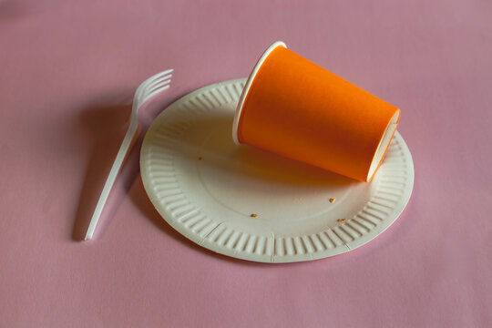 Orange disposable cup lying on a disposable plate with crumbs after food, next to a disposable fork on a pink background.