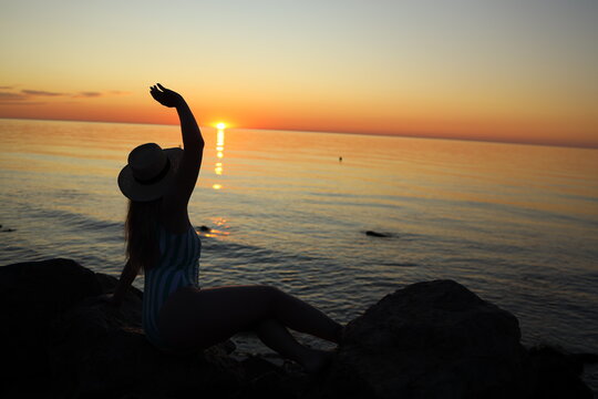 Woman waves goodbye to the sun while sitting on rocks by the sea during sunset at the beach