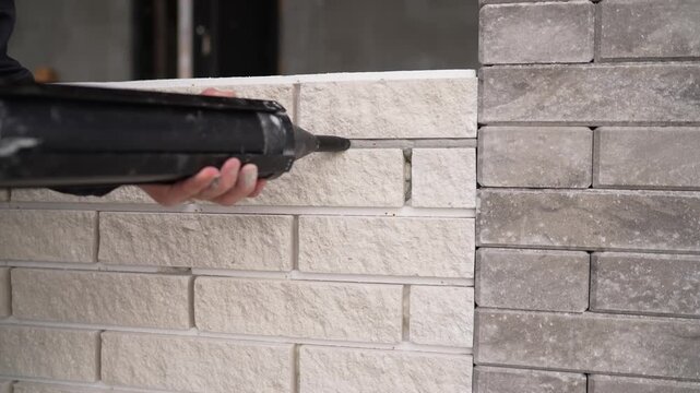 Close up of a builder applying grout to a decorative brick wall. Worker uses a pointing gun for precision work on the facade. Neutral colors and an industrial mood, ideal for construction themes.