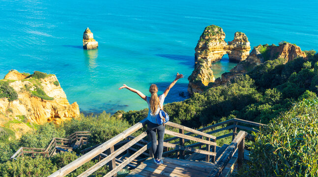 Empowered woman with open arms celebrating freedom at Praia do Camilo cliffs, Lagos, Algarve, Portugal - Success and travel bliss concept.