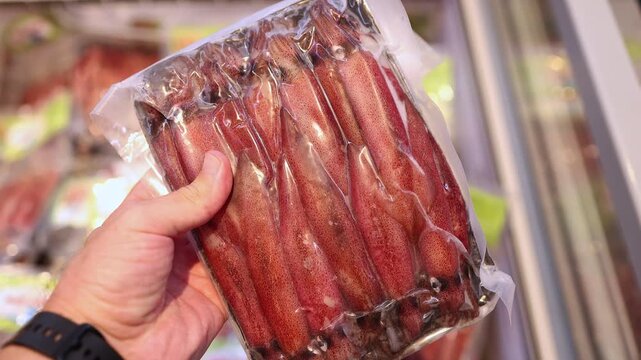 Close up of a hand holding a vacuum pack of fresh raw whole squid in a supermarket. Customer choosing seafood product in a store. Frozen cephalopod mollusk in plastic package.