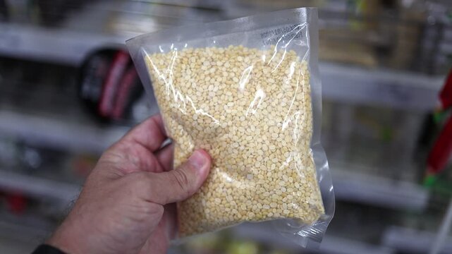 Close up of a hand holding a plastic pack of dry yellow split lentil in a supermarket. Customer choosing organic dal pulse seed in a grocery store. Healthy nutrition and vegan protein concept.