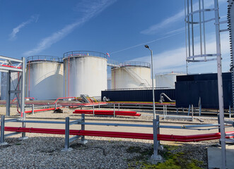 Obraz premium Industrial fuel storage tank farm with large white cylindrical oil and diesel tanks, red pipelines, and infrastructure at a petroleum reserve facility in the Netherlands