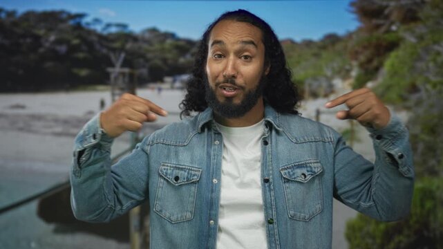 Man in denim jacket and white t shirt points finger to chest on a sunny beach boardwalk, smiling with relaxed hands and open stance; confidence.