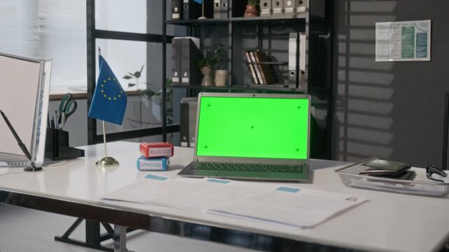 Medium full shot of laptop with green chromakey screen, next to visa applications, approved and rejected stamps, European Union flag, on consular officer desk at foreign embassy or diplomatic mission