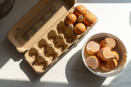 Brown eggs in cardboard carton with cracked egg shells in bowl overhead