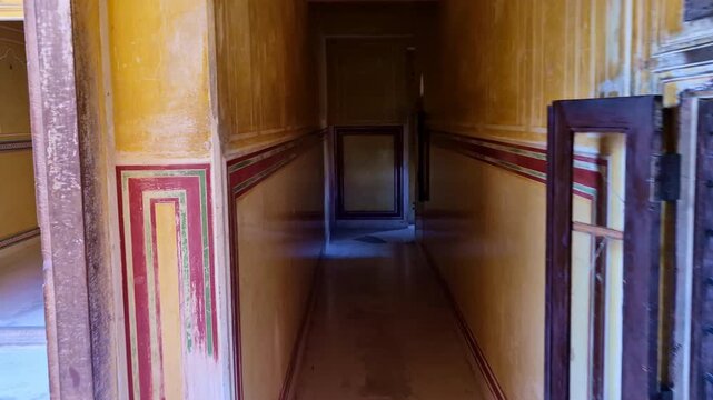 A narrow historic corridor inside an old fort or palace, with yellow plastered walls, red-green painted border lines, small arched window openings with wooden shutters, and bright sunlight entering.