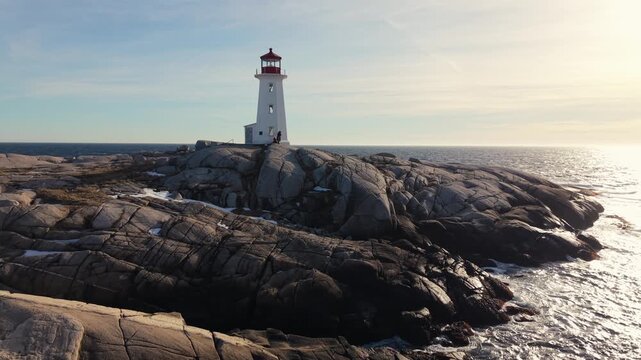 Aerial drone footage sunset: snow-covered Peggy's Cove lighthouse on Atlantic coastline near Halifax. Nova Scotia Canada winter wonderland, warm golden light over ocean 4K.