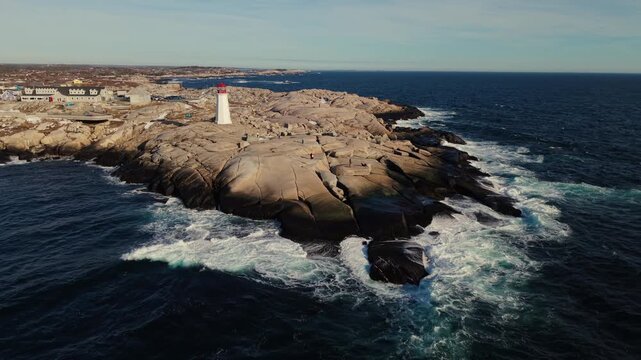 Drone aerial shot: Peggy's Cove lighthouse and snowy granite rocks at sunset over Atlantic Ocean. Halifax suburb Nova Scotia Canada, dramatic winter seascape with golden light 4K.