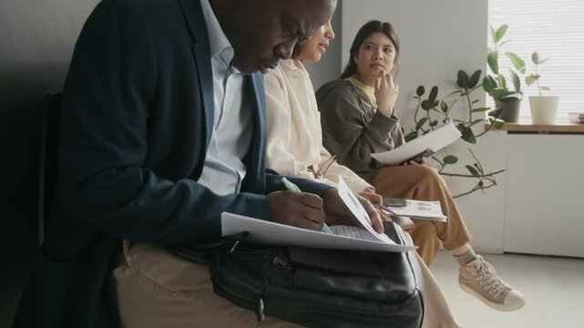 Side medium close-up of African American businessman in suit sitting on chair while waiting in queue at embassy, filling in visa application form, answering questions while checking passport number