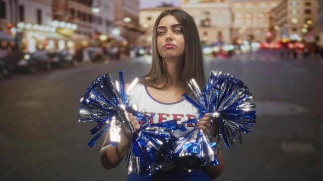 Woman cheerleader holding blue metallic pompoms with both hands, gazing upward while standing in a city street lined with lights at dusk; contemplation.