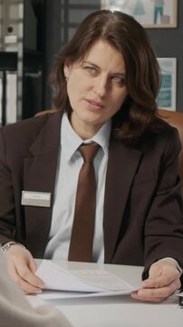 Vertical medium shot of polite mature Caucasian female consular officer talking to anonymous visa applicant during interview at embassy, examining form and asking questions to verify information