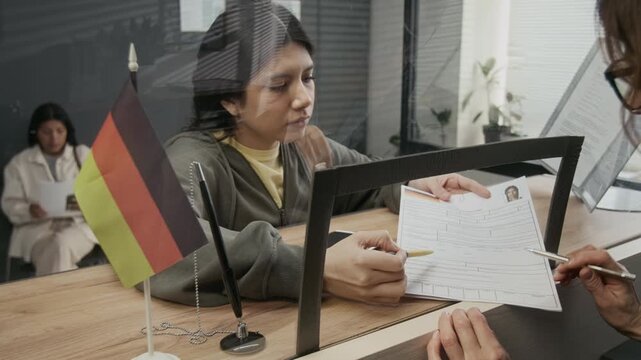 Medium close-up of young Hispanic girl and Caucasian female immigration authority officer clarifying personal details and purpose of visit on application form while obtaining German student visa