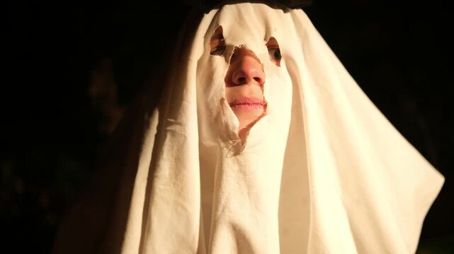 Funny scene of a person wearing a white sheet with eye and mouth holes for a ghost costume, making scary faces and noises at night during a fun halloween celebration party in the dark