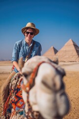 Vacation travel trip journey in holiday season. A man riding a camel in front of the majestic pyramids of Giza. He wears a hat, sunglasses, and a shirt.