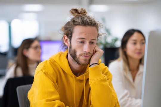 Young caucasian male adult frustrated bored at computer in open office workspace wearing yellow hoodie