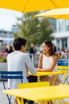 Young asian female and young asian male adult chatting at yellow outdoor cafe table under umbrella in sunny urban parklet