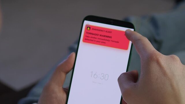 User receives an urgent Tornado Warning emergency alert on a smartphone. A finger taps the red notification. Perfect for disaster preparedness, safety training, and weather-related content. High