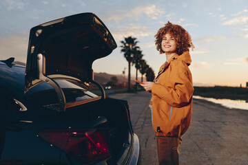 Fototapeta premium Smiling woman with curly hair wearing an orange jacket stands near an open car trunk holding a smartphone on a sunny road lined with palm trees. Lifestyle and travel concept.