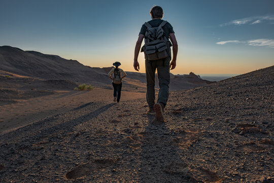 ouple of hikers with backpacks walking towards the sunset in a rocky desert. Following footprints, partnership and outdoor adventure travel concept in scenic wilderness.