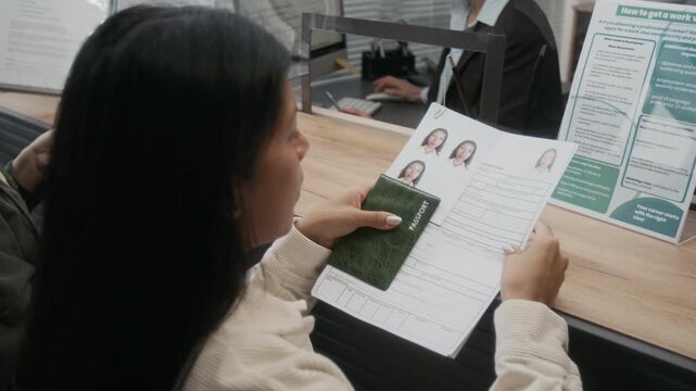 Over-shoulder shot of mature Hispanic woman standing by counter, carefully checking personal details in application form, while daughter is talking to visa center employee for assistance