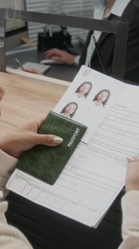 Vertical close-up of hands of anonymous woman checking personal information in application form, holding passport and photos, while standing in queue at counter at visa center