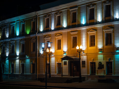 Elegant architectural facade of historic multi story building featuring symmetrical white pilasters and yellow walls beautifully illuminated by multicolored blue and warm yellow spotlights