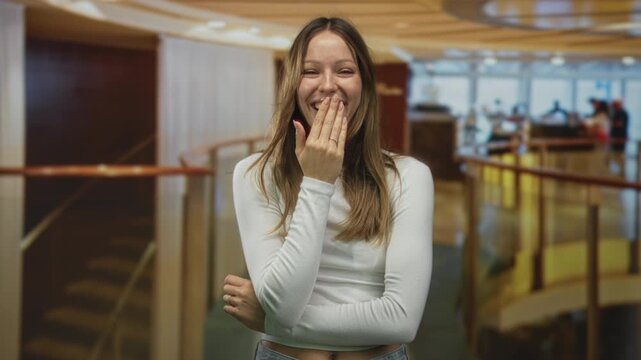 Woman laughing with hand covering mouth inside hotel lobby with curved railing and staircase visible; playful shyness joy.