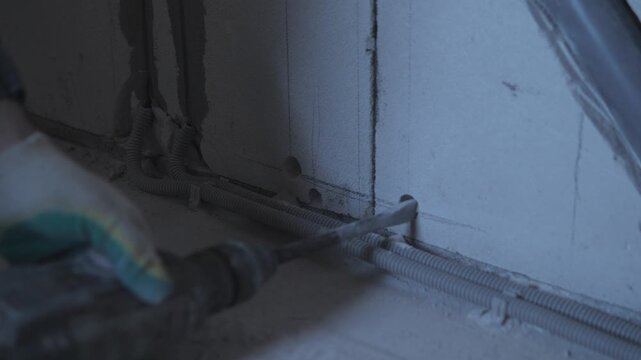 Hand Placing Flexible Conduit Along Wall Base, Closeup Of Gloved Hand Guiding Corrugated Pipe Into Drilled Holes For Wiring, Indoor Renovation Scene, Measured Positioning And Secure Fitting
