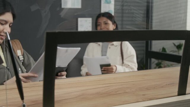 Medium panning shot of anonymous female consular officer handing documents and passport with approval stamp to young Hispanic woman, hugging waiting friend, happy at being granted tourist visas