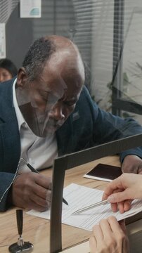 Vertical medium close-up of mature African American businessman completing visa application with assistance from anonymous embassy officer, pointing to questions and signatures on form