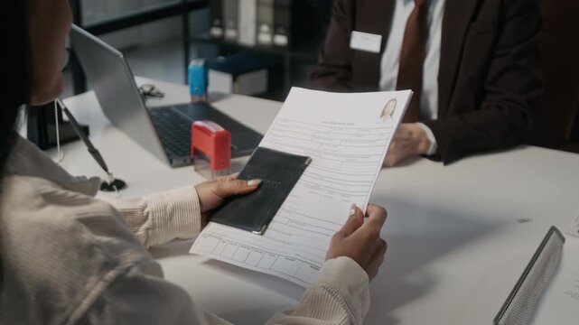 Medium shot of polite mature Caucasian female embassy immigration officer asking Hispanic woman questions on bank statement, while conducting interview on travel visa application