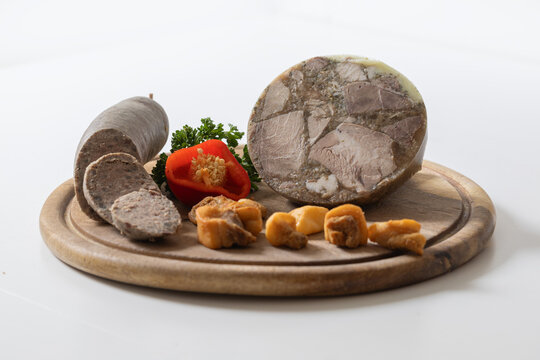 Traditional Czech pork delicacies: sliced liver sausage, headcheese, and pork rinds on a board with red pepper and parsley, white background.