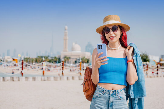 Young woman smiling, wearing a straw hat and sunglasses, holding a phone on a sandy beach with the Dubai city skyline in background