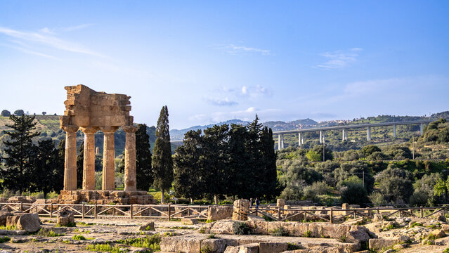 Ancient Temple of Castor and Pollux in the Valley of the Temples in Agrigento, Sicily, with a modern elevated highway (viaduct) stretching across the landscape in the background.