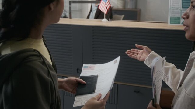 Over-shoulder follow shot of two young and mature Hispanic women checking application forms, then walking over to counter and submitting documents to Caucasian USA consulate officer