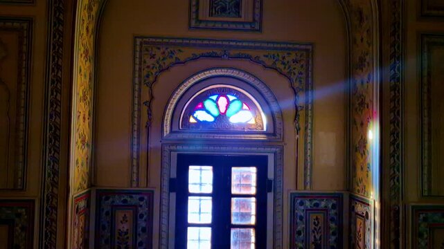 Ornate arched wall in a Jaipur heritage palace, with delicate floral frescoes, carved borders, and a glowing stained-glass window panel reflecting traditional Rajasthani royal design.