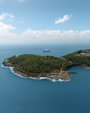 Aerial view of the lush tropical vegetation and rocky coastline of the Salvation Islands with a ship anchored in the blue sea at Iles du salut, French Guiana.